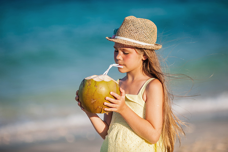 kleines Mädchen, das am Strand aus einer Kokosnuss trinkt.