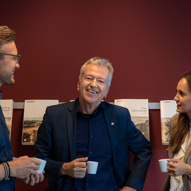 Drei lächelnde Personen halten Kaffeetassen in der Hand und geniessen einen fröhlichen Moment zusammen.