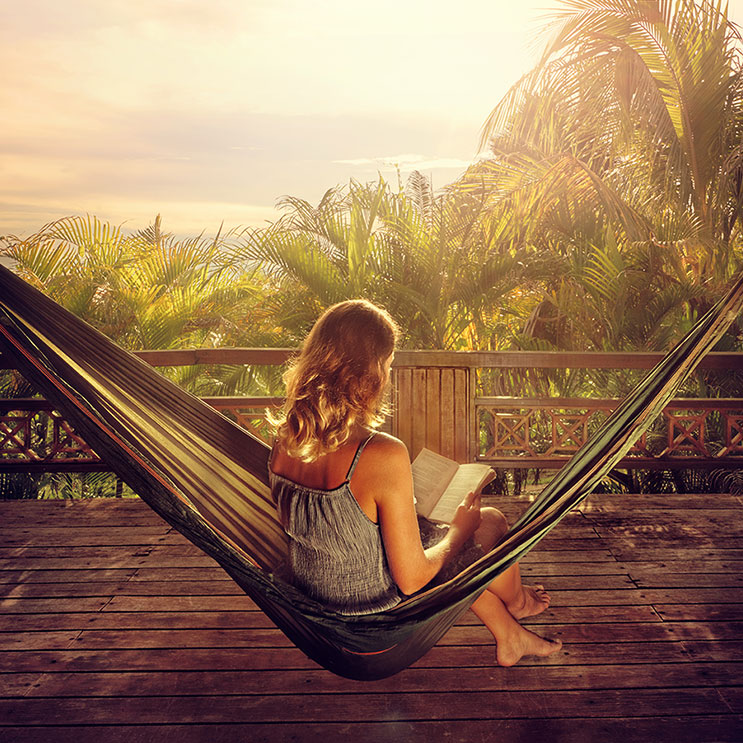 Une femme se détend dans un hamac et lit un livre, entourée de nature et de calme.